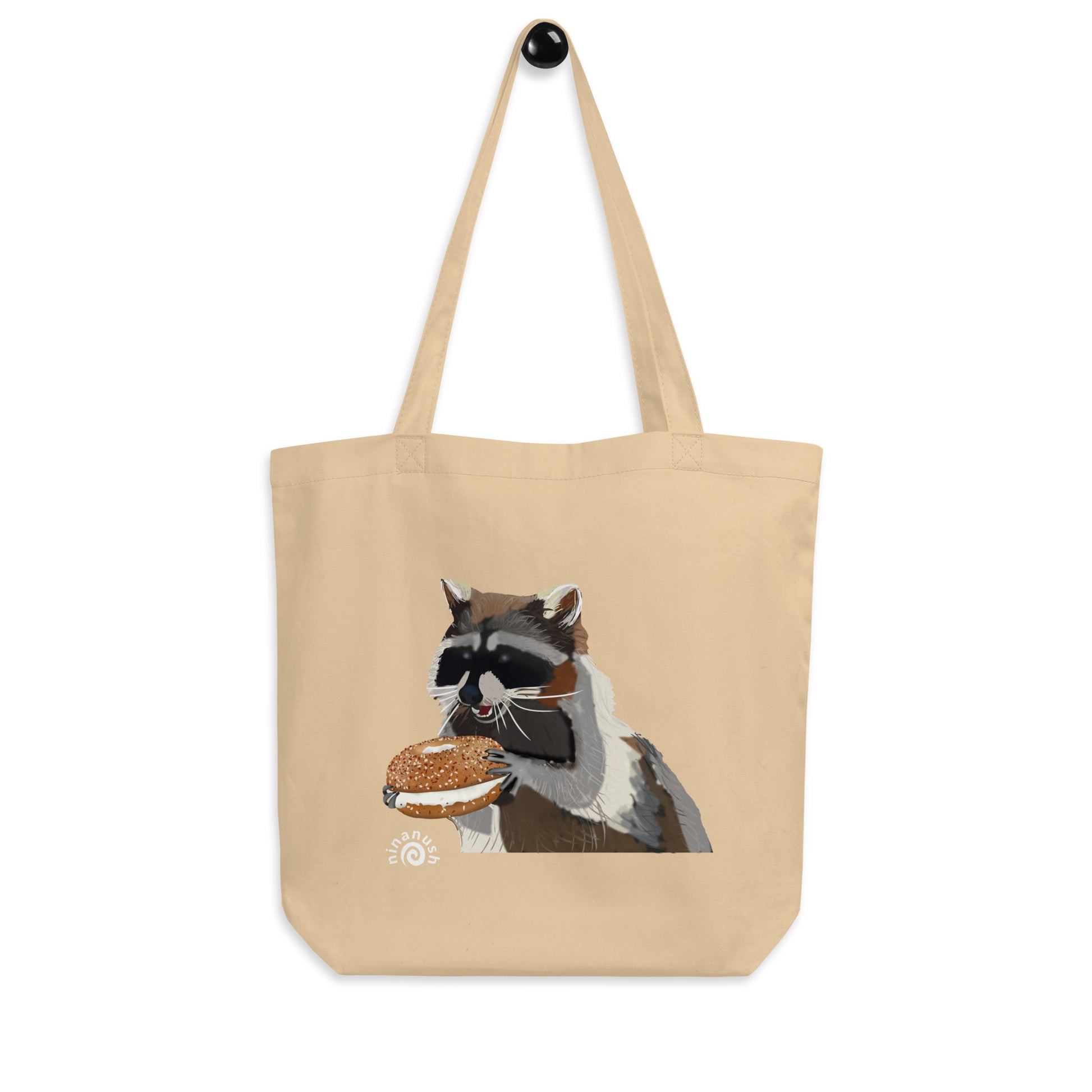 Beige tote bag with funny raccoon design and everything bagel - This everything bagel tote bag with a raccoon eating bagel is perfect for bagel lovers, foodies and raccoon enthusiasts. The funny foodie design is totally unique and the organic cotton tote bag is perfect for everyday adventures. Carry your groceries, bagels, books and everything in between in this sustainable shopping tote bag. 